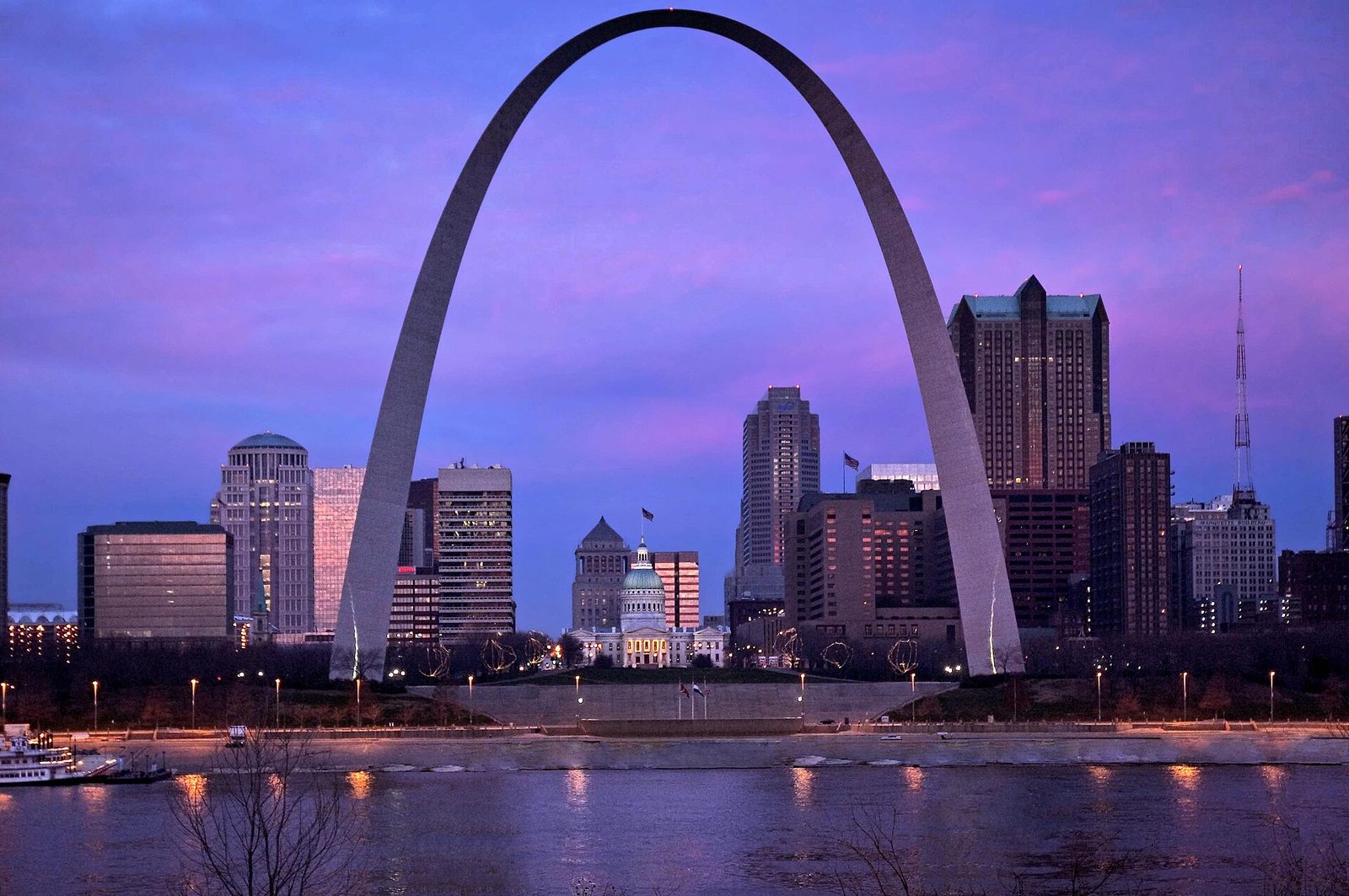 The Gateway Arch and the St. Louis riverfront at dawn, seen from across the Mississippi River with the sky turning orange behind the skyline.